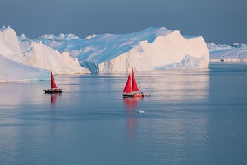 Little red sailboat cruising among floating icebergs in Disko Bay glacier during midnight sun...