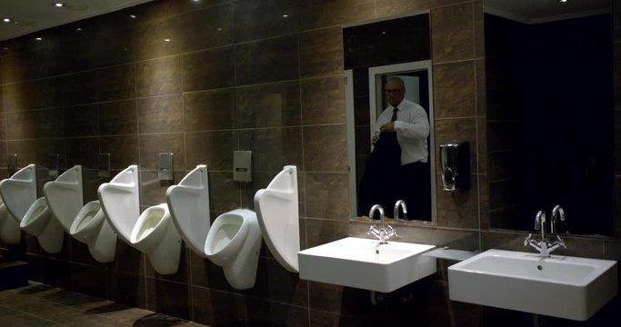 A business man comes out of the toilet cubicle into the bathroom and puts on a suit jacket