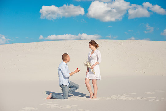 Young man stands on one knee proposes to marry to his lover on a background of white sand, dune. Love story in the void.