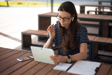 Girl, student, using the tablet, working online, using voice messages. Near notebook, a smartphone, wireless headphones.