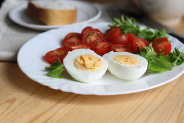 Healthy and tasty breakfast, boiled egg, cut in half, red ripe cherry tomatoes and parsley on a white plate, a piece of bread on a saucer on a napkin, fork, knife, cup, wooden table