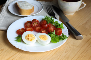 Healthy and tasty breakfast, boiled egg, cut in half, red ripe cherry tomatoes and parsley on a white plate, a piece of bread on a saucer on a napkin, fork, knife, cup, wooden table