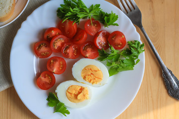 Healthy and tasty breakfast, boiled egg, cut in half, red ripe cherry tomatoes and parsley on a white plate, a piece of bread on a saucer on a napkin, fork,  wooden table, top view