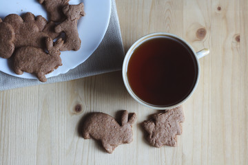 Sweet breakfast, tea break, sweet snack, cup of tea, homemade cookies in the form of a bunny and christmas tree on a wooden table, a white plate with cookies on a napkin, top view