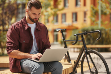 Modern blogger. Portrait of young man with stubble in casual clothes working on laptop while sitting on the bench near his bicycle