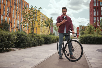 Chatting with friend. Full length of handsome man with stubble in casual clothes and eyeglasses holding mobile phone while walking with his bicycle outdoors