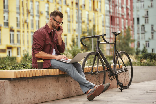 Important talk with client. Handsome and young man with stubble in casual clothes working on laptop and talking by phone while sitting on the bench near his bicycle