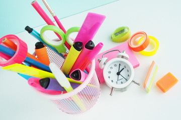 school stationery: felt-tip pens, rulers, pens, pencils and scissors in a mesh basket, multi-colored neon scotch tape, sharpener and white alarm clock on pink stickers...
