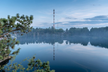 Fototapeta na wymiar Dramatic scenic view through the trees of the granite quarry filled with blue water, and the opposite shore overgrown with trees after sunset at dusk in the summer. High tower on the opposite bank. 