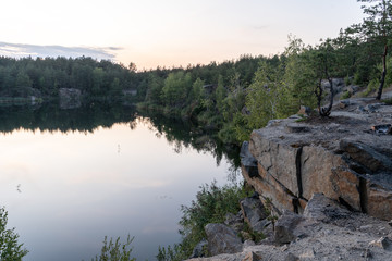 Dramatic scenic panorama of the granite quarry filled with water, and the opposite rocky shore overgrown with after sunset at dusk in the summer. 