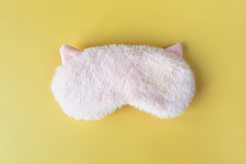Pastel pink fluffy fur sleep mask with small ears on pastel yellow paper background. Top view, flat lay. Concept of vivid dreams. Accessories for girls and young women