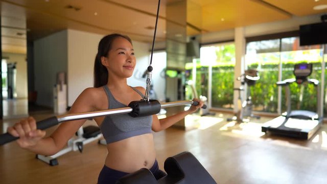 Strong fit Asian woman works out her​ lats at the pulldown bar in a resort gym.