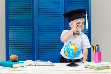 Clever and smart child, prodigy. A little toddler boy in a graduation cap is examines a globe through a magnifying glass. Sitting at a desk with books on blue wooden wall background