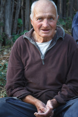 Close-up portrait of a very old man with arms crossed, sits on the ground on a blurred background of wood fence, selective focus