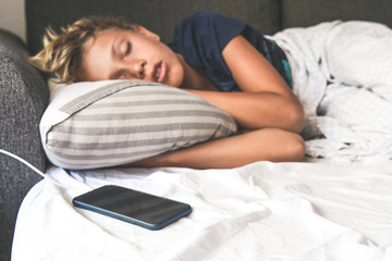 Young boy sleeping with hands under the pillow near the cellphone. Beautiful child sleeps alone in the bed dreaming about online games. Technology addicted, youth, new generation trend concept