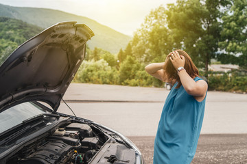 Young woman tourist driver standing by the road by the car with the hood open dead engine broken automobile malfunction repair in a summer day holding her head in trouble