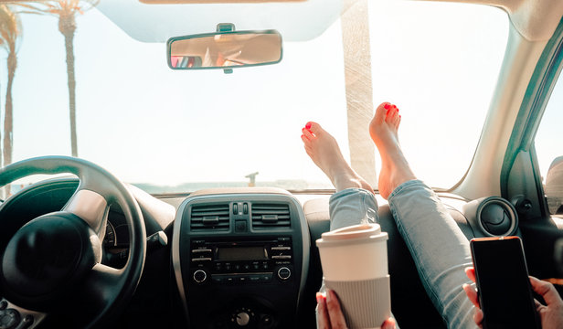 Woman drinking cappuccino inside car with feet on dashboard - Girl relaxing in auto looking cellular with palms clear sky in background  - Traveler concept - Focus on feet - Image