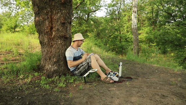young handsome caucasian naturalist scientist sits in a green forest under a tree and makes notes in his notebook, medium plan