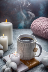 Fototapeta na wymiar cup of hot drink in white knitted cover placed on the book candles and cotton around hugge style