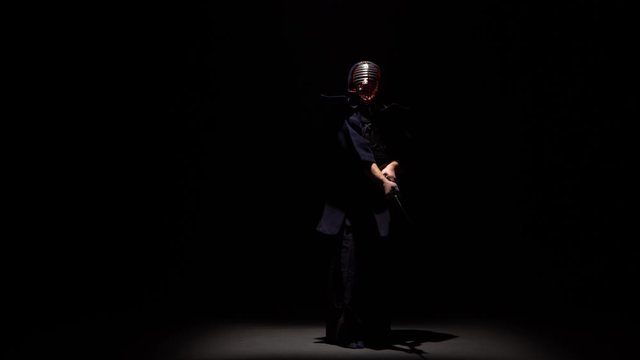 Kendo fighter performing martial art with Katana sword at dark studio under spotlight.