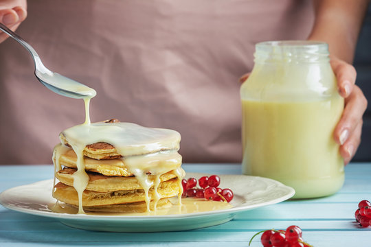 A girl in a pink apron adds condensed milk to pancakes. Delicious healthy food. Delicious pancakes with sauce and red currants. Close-up photograph of fresh natural dessert.