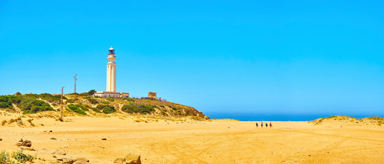 Tourists walking in direction to Faro de Trafalgar Beach, a broad beach of The Cabo de Trafalgar Cape Natural Park with the famous Lighthouse in the background. Barbate, Los Caños de Meca, Cadiz. Anda