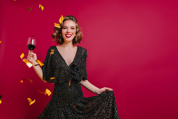 Blissful female model with shiny curly hair posing with wineglass on claret background. Emotional...