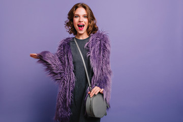 Lovely young female woman with trendy gray handbag posing on purple background with surprised...