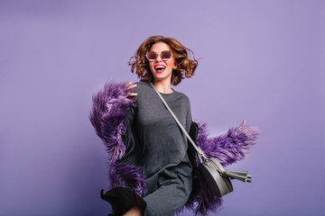 Attractive girl in gray dress and black sunglasses dancing in studio on purple background. Indoor portrait of charming young woman in fur coat having fun during photoshoot.