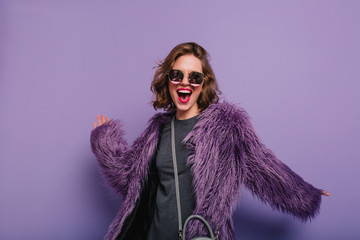 Inspired lady with stylish makeup dancing in purple fur coat and laughing. Indoor portrait of glad lovely woman in soft jacket and dark sunglasses.