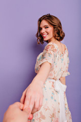 Fototapeta na wymiar Portrait from back of laughing romantic girl holding photographer's hand. Studio shot of carefree young lady in pink summer dress posing on purple background.