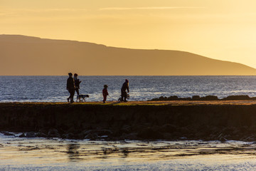 A family walking on a Salthill, Galway, Ireland jetty at sunset appear in silhouette against a...