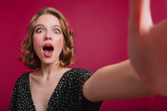 Surprised brunette woman with little tattoo on arm making selfie. Indoor portrait of amazed female model taking picture of herself in bright background.