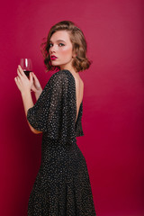 Sad woman in elegant long dress looking over shoulder, holding wineglass. Charming pensive female model with short haircut enjoying wine at party.