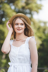 front view of beautiful young girl in white dress touching straw hat while smiling and looking away
