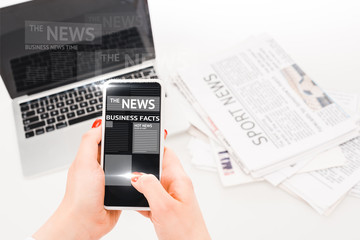 selective focus of woman holding smartphone with business news illustration near laptop and newspapers