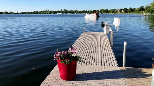 US flags and red flowers in pot on a boat dock while a pontoon on a beautiful Minnesota lake passes by on the Fourth of July.