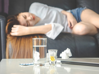 sickness and gynecology concept from abstract blurred of woman with allergy, feeling dizzy and fever sleep on sofa in home. focus at pills and glass of water on the table.