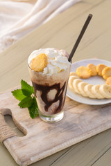 Tasty milkshake drink in glass mug with plastic tube and slice of banana on chopping board on wood table