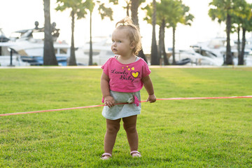 A little girl of 1 year old running on a green meadow in a park among palm trees and cutting a red ribbon