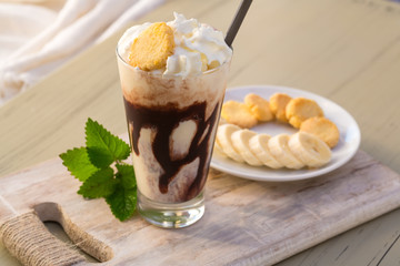 Tasty milkshake drink in glass mug with plastic tube and slice of banana on chopping board on wood table