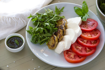 Caprese salad with mozzarella, tomato, basil and grilled eggplant on white plate. Top view on background