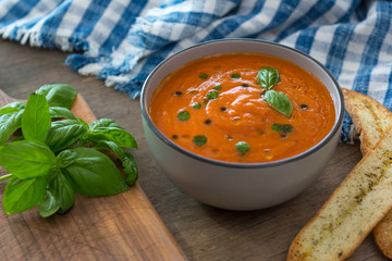 A bowl of fresh tomato soup in white ceramic bowl, garnished with basil, croutons, seasoning and a drizzle of olive oil, and served with crusty bread.