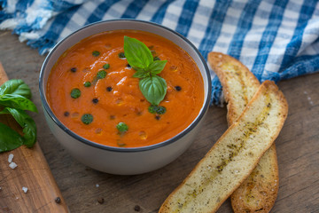 A bowl of fresh tomato soup in white ceramic bowl, garnished with basil, croutons, seasoning and a drizzle of olive oil, and served with crusty bread.