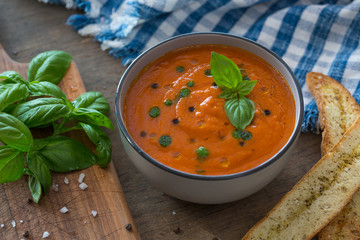 A bowl of fresh tomato soup in white ceramic bowl, garnished with basil, croutons, seasoning and a drizzle of olive oil, and served with crusty bread.