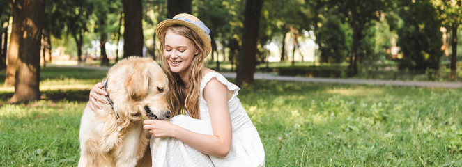 panoramic shot of beautiful girl in white dress and straw hat petting golden retriever while sitting on meadow
