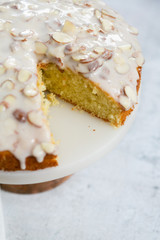slice of almond olive oil cake with brown butter icing and sliced almonds, dripping icing on cake, marble and wood cake stand, styled bakery