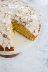 slice of almond olive oil cake with brown butter icing and sliced almonds, dripping icing on cake, marble and wood cake stand, styled bakery