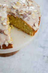 slice of almond olive oil cake with brown butter icing and sliced almonds, dripping icing on cake, marble and wood cake stand, styled bakery
