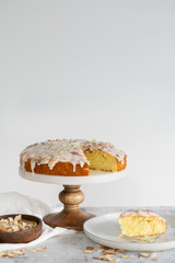slice of almond olive oil cake with brown butter icing and sliced almonds, dripping icing on cake, marble and wood cake stand, styled bakery